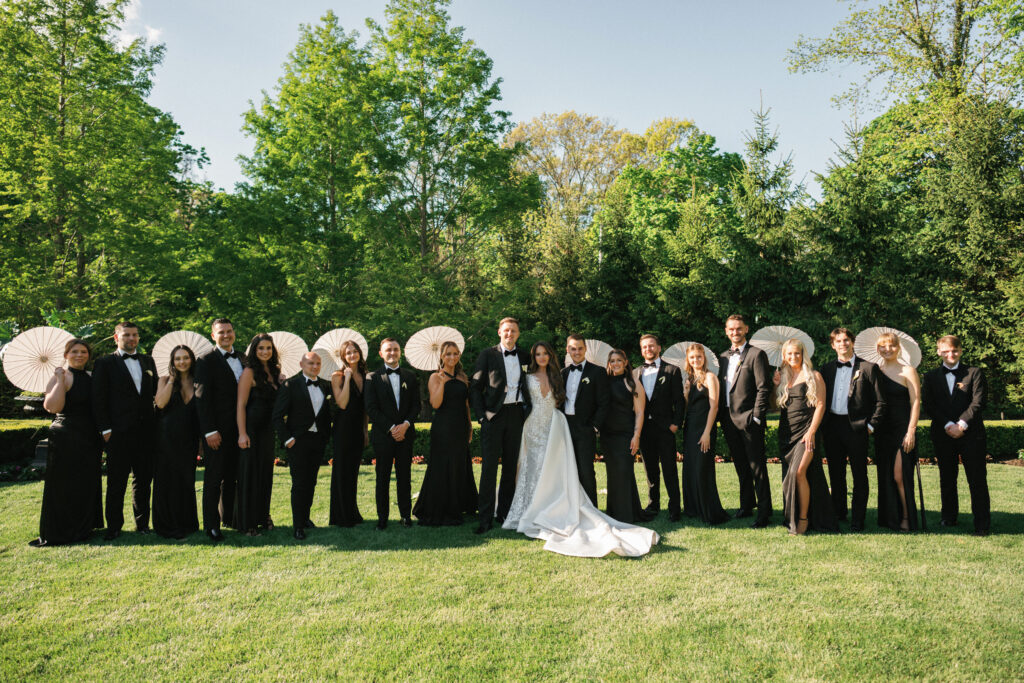 Vogue-style black tie wedding group photo outdoors, showcasing a sophisticated modern bride and groom with their large family and friends surrounded by lush green trees.
