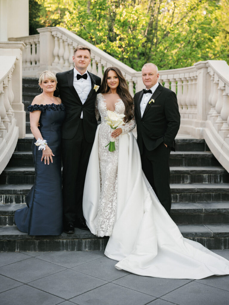 High-end family wedding portrait on a grand stone staircase of a luxury estate, featuring a bride in a couture lace gown with a dramatic overskirt posing with the groom and parents.
