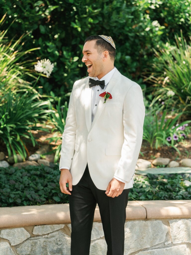 A laughing groom in a white tuxedo jacket and a traditional yarmulke turning around to react to his bride during a first look photo and film session.