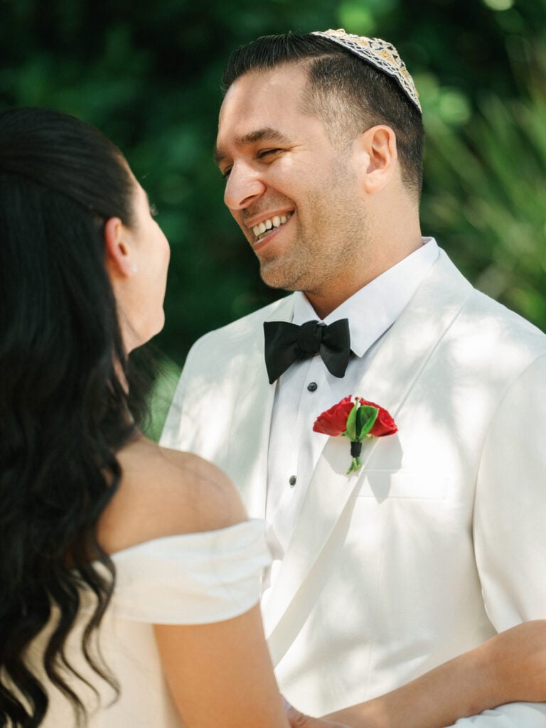 Close-up portrait of a bride with long dark hair and a groom in a white suit smiling and looking at each other after their wedding first look reveal.