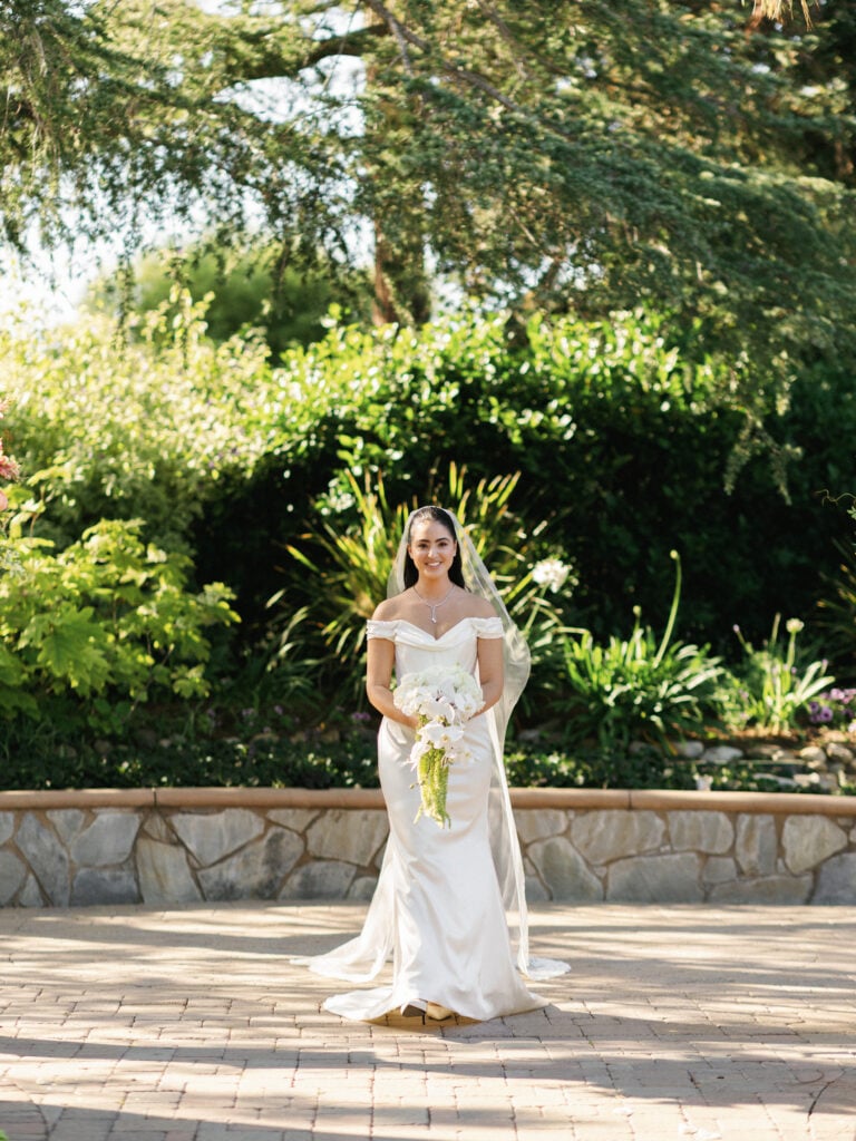 A smiling bride in an off-the-shoulder white dress holding a bouquet as she walks up for a pre-ceremony first look reveal session outdoors.