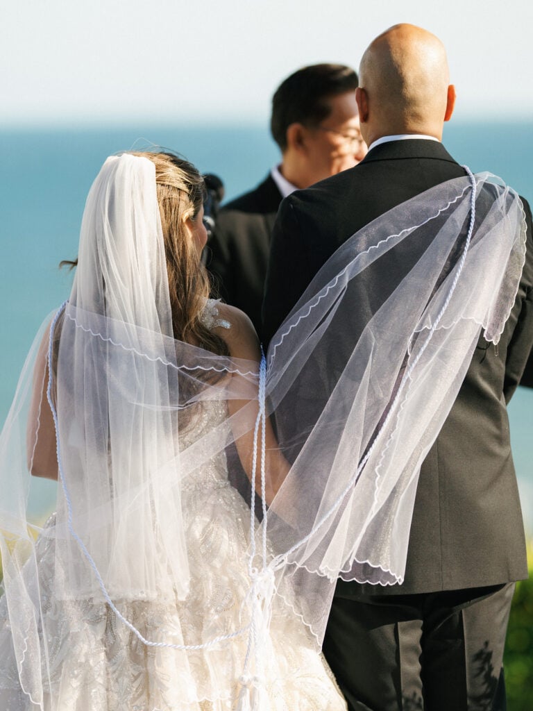Rear view of a bride and groom standing at the altar with a sheer wedding veil draped over the groom's shoulder during an outdoor ceremony at The Ritz-Carlton, Laguna Niguel.