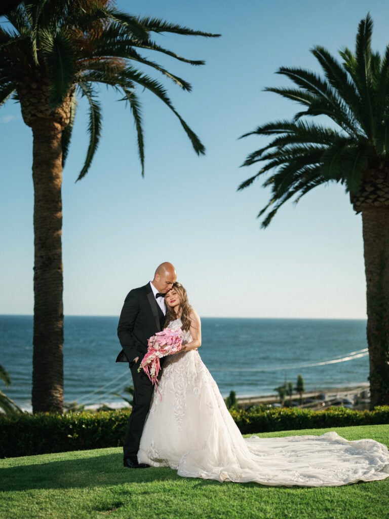 Full-length wedding portrait of a bride leaning her head on the groom's shoulder on a green lawn between two palm trees overlooking the ocean at The Ritz-Carlton, Laguna Niguel.