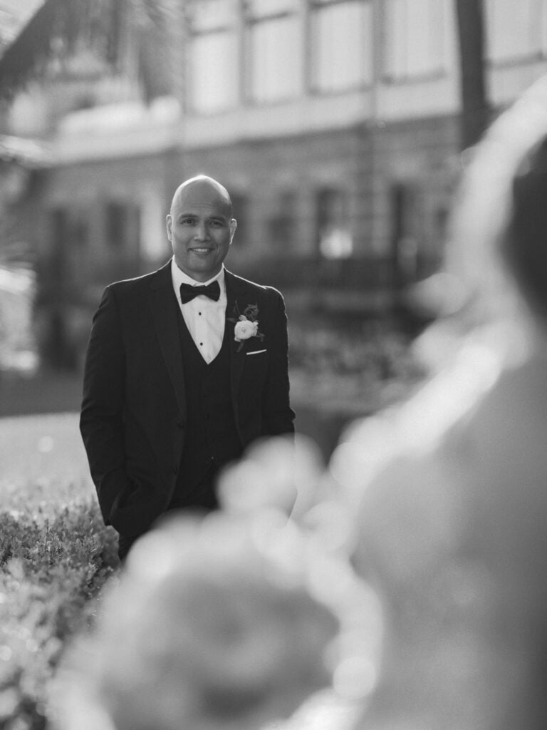 Black and white over-the-shoulder shot of a smiling groom in a three-piece tuxedo with hands in pockets, looking at his bride during a first look wedding reveal.