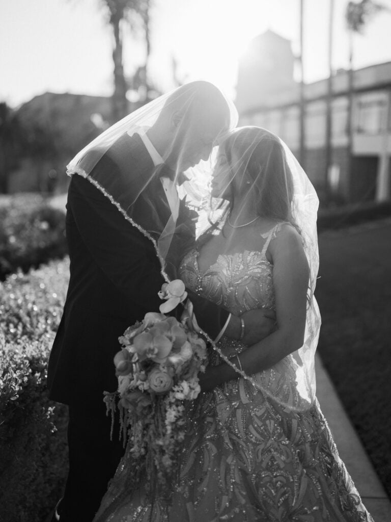 Black and white portrait of a groom in a tuxedo and a bride in a heavily embellished gown touching foreheads under a veil during a backlit golden hour first look session.