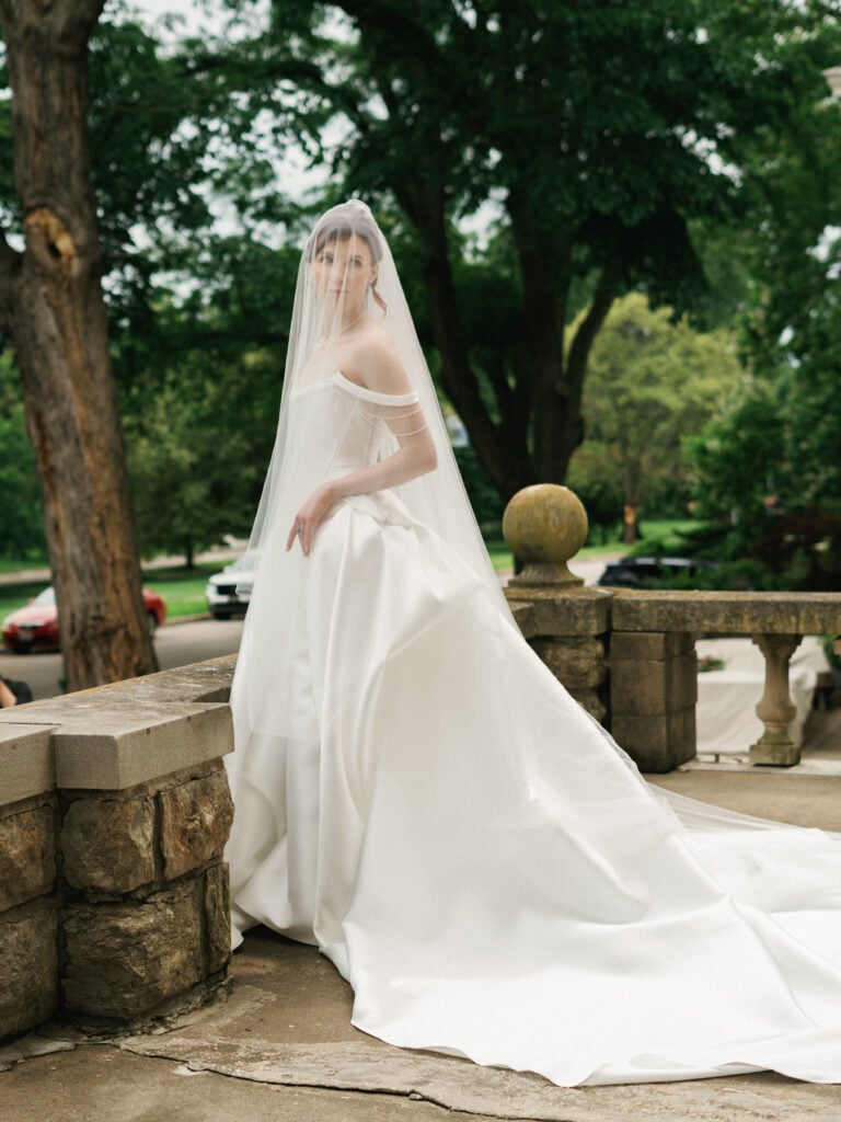 Bride standing in profile on a stone terrace with her head turned to the camera, wearing a voluminous satin wedding gown with a long train and a sheer cathedral veil covering her head and shoulders.