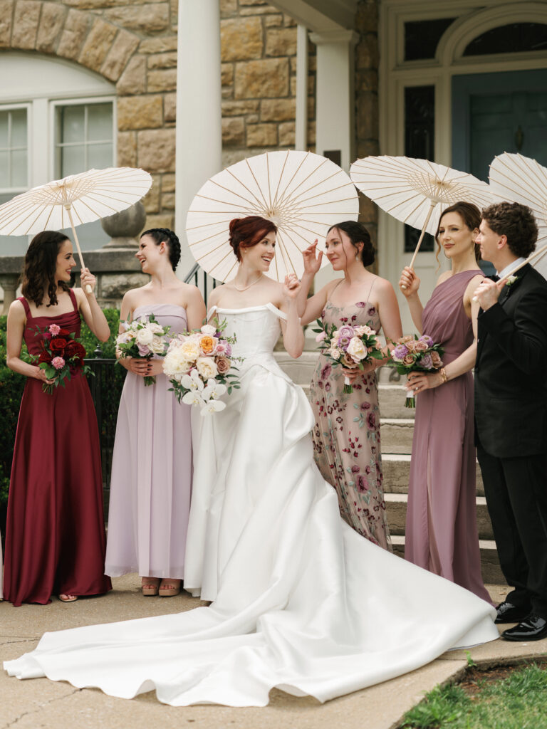 Bride in a dramatic structural gown posing with bridesmaids in mismatched burgundy, lavender, and floral print dresses holding white parasols.