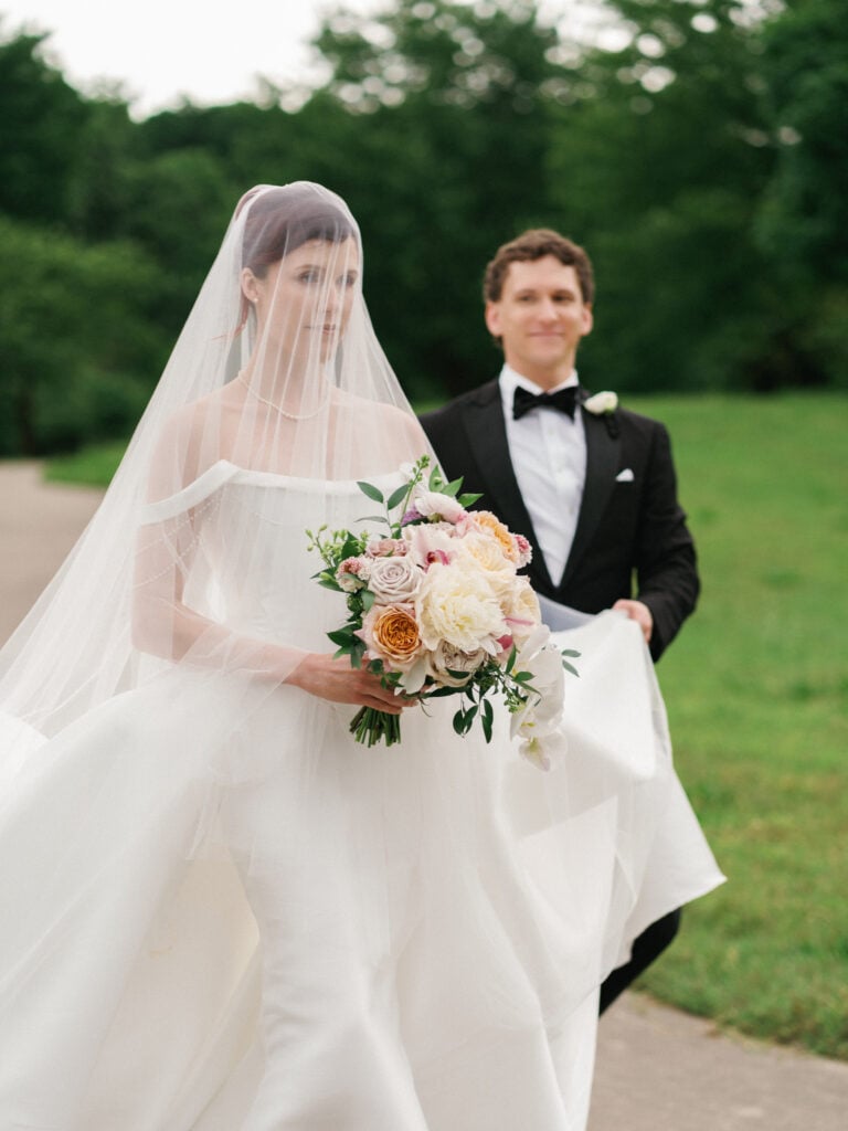 Bride walking forward with her face covered by a sheer wedding veil, while the groom walks behind her holding up the train of her white gown on a path.