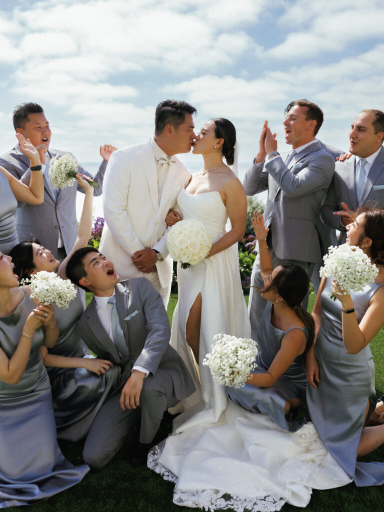 Vertical close-up of a bride and groom kissing on a lawn, surrounded by bridesmaids kneeling in dusty blue satin dresses and groomsmen in grey suits cheering.