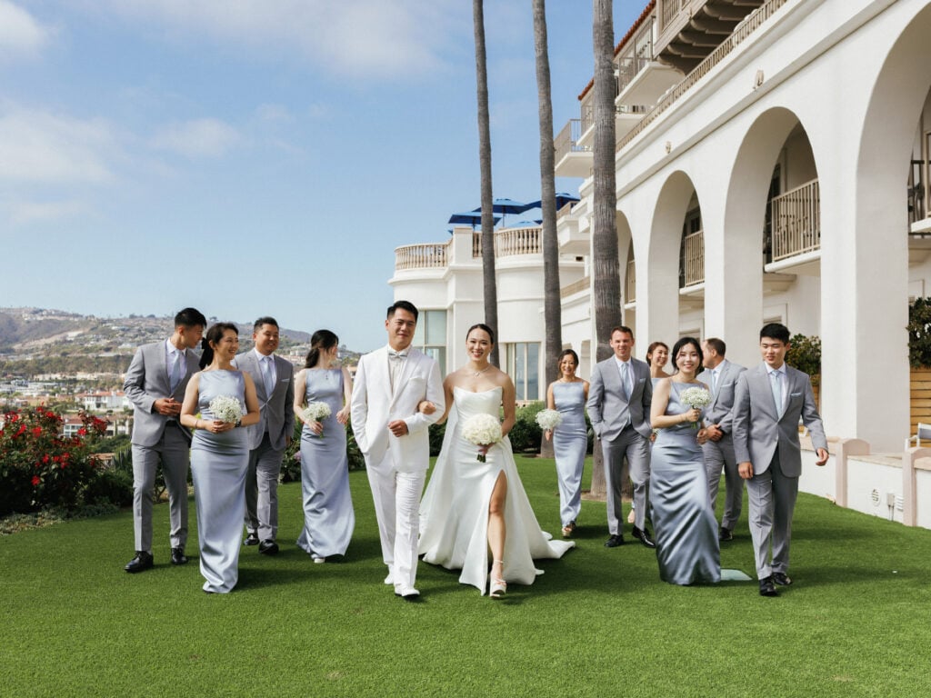 Bride in a strapless dress with a high slit and groom in an all-white tuxedo holding hands and walking forward on a green lawn, flanked by bridesmaids in dusty blue satin dresses and groomsmen in grey suits next to a white arched building with tall palm trees at The Ritz-Carlton, Laguna Niguel.
