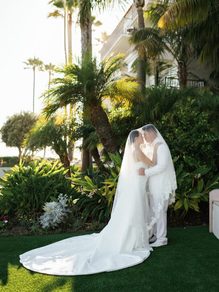 Groom in an all-white tuxedo and bride in a strapless wedding dress covered by a lace-trimmed cathedral veil under palm trees.