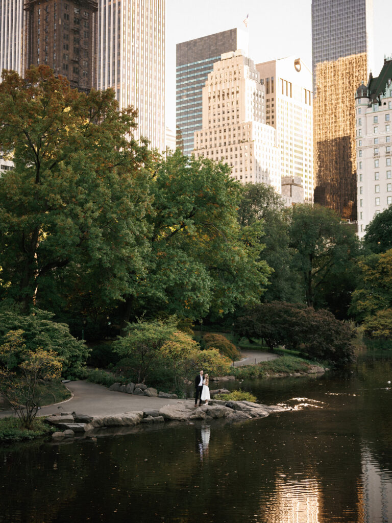 A wide wedding portrait of a couple by the Central Park pond with The Plaza Hotel and Midtown Manhattan skyscrapers behind them.