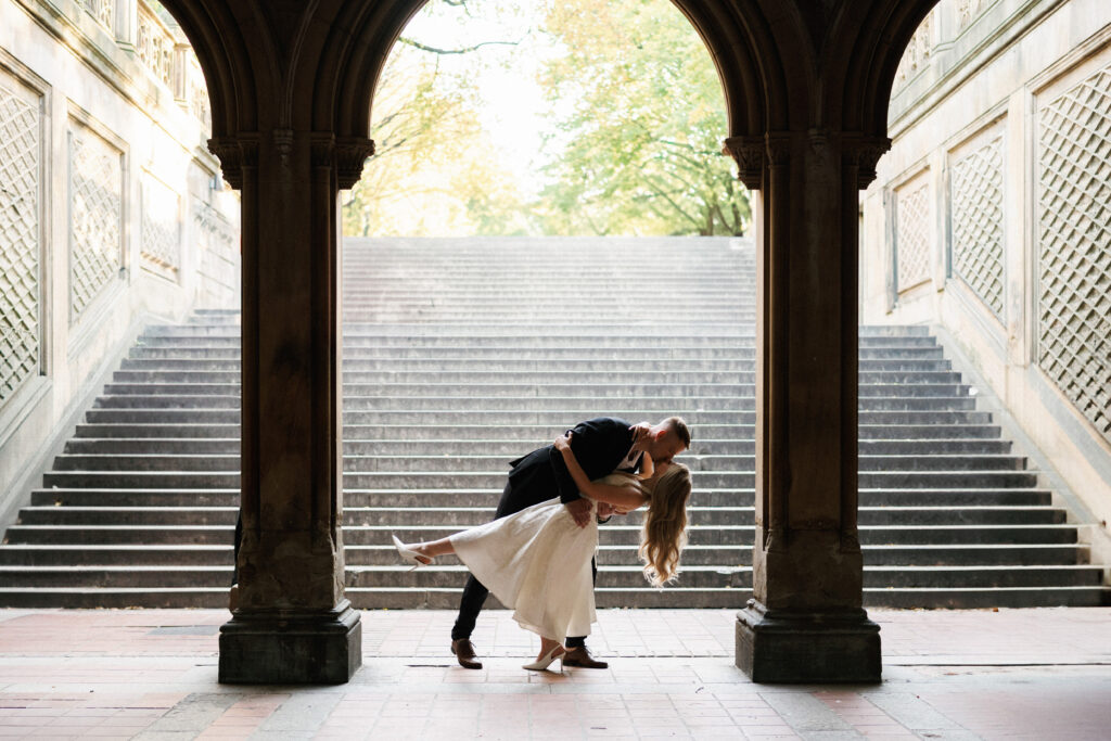 A groom dips and kisses his bride inside the historic Bethesda Terrace arcade with grand stone stairs in Central Park.