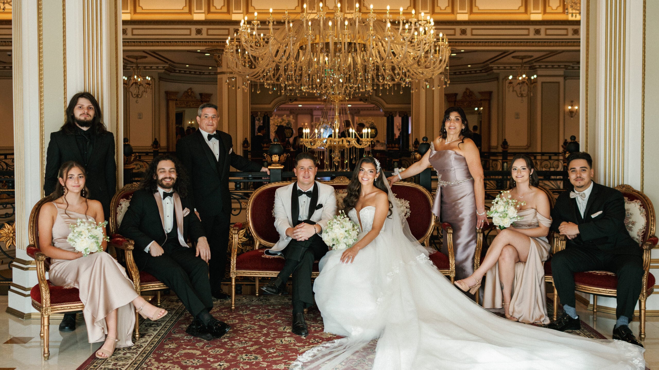 Royal-inspired family wedding portrait inside a luxury historic hotel ballroom, featuring the bride and groom on a vintage velvet sofa under a grand crystal chandelier.