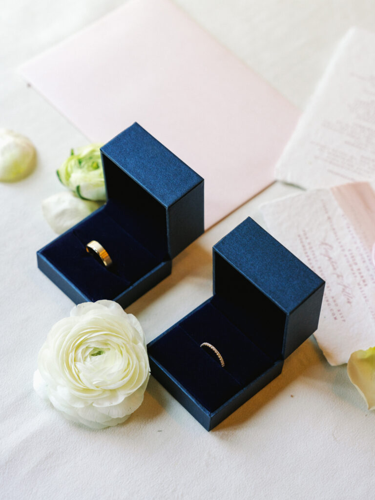 Gold wedding bands resting inside two open navy blue velvet boxes next to a white ranunculus flower and blush pink invitation cards for elegant wedding detail photos.