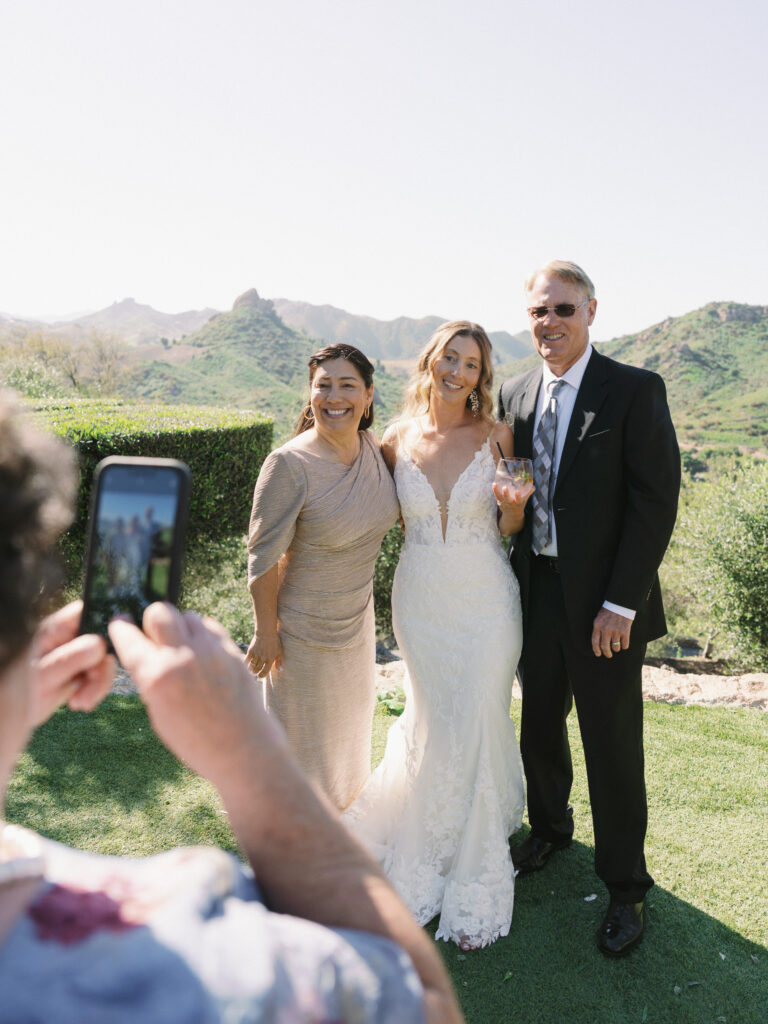 Guest taking a phone photo of a modern bride in a lace gown with her parents on a scenic overlook lawn with a Malibu mountain backdrop.