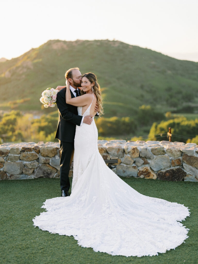 Romantic Malibu wedding photography of a newlywed couple embracing on a stone overlook during a scenic golden hour sunset.