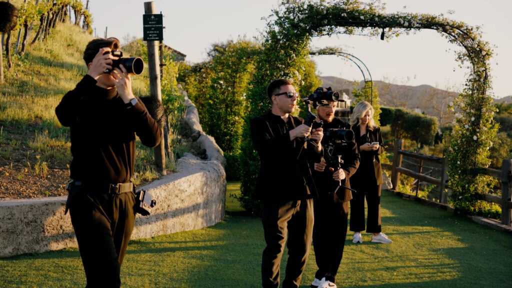 A full luxury wedding media production crew, including photographers and videographers with gimbals, filming at a scenic vineyard during golden hour.