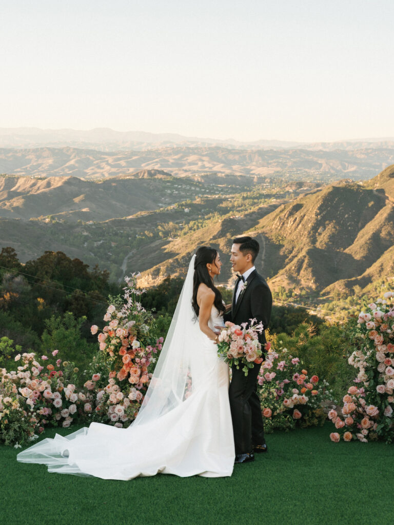 Bride and groom posing during sunset at a mountain overlook ceremony venue in Topanga California, surrounded by lush peach and pink rose floral installations.