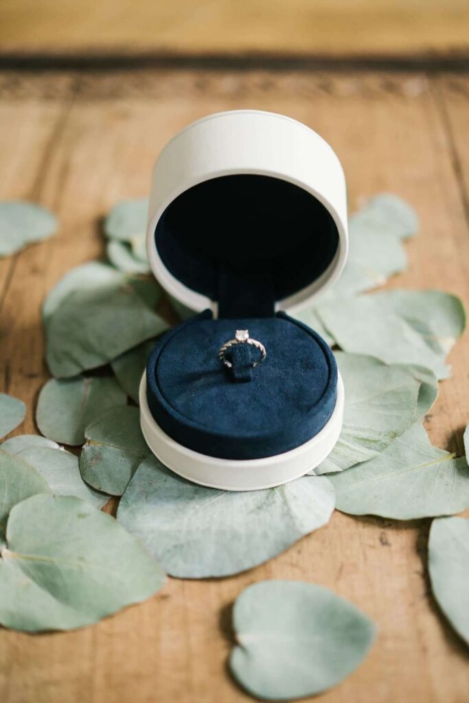 Solitaire diamond engagement ring inside a round navy blue and white velvet box, arranged on a bed of green eucalyptus leaves for bridal wedding detail photos.