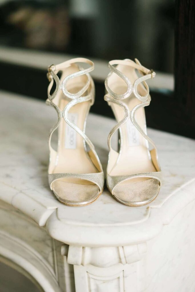 Gold metallic strappy Jimmy Choo wedding sandals standing on a classic white carved marble fireplace mantel during bridal prep.