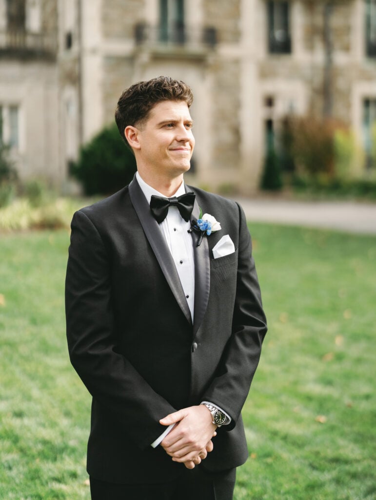 Groom in a black tuxedo standing on a lawn during a wedding first look reveal session, smiling and waiting for the bride in front of a stone estate.