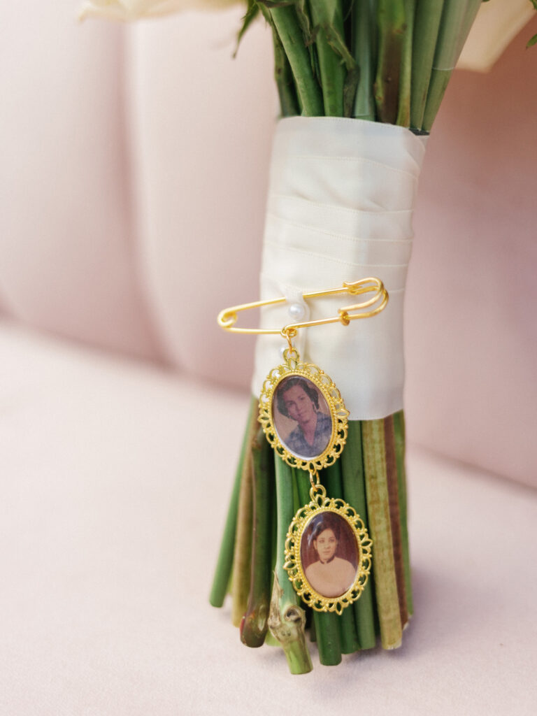 Custom gold bridal bouquet charms with oval photo portraits honoring deceased family members, pinned to a white silk ribbon wrapped around the flower stems.