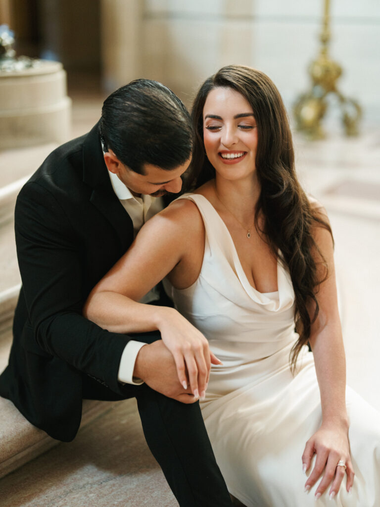 Editorial black-tie couple portrait highlighting a minimalist cowl-neck bridal dress against the neoclassical limestone walls of San Francisco City Hall.
