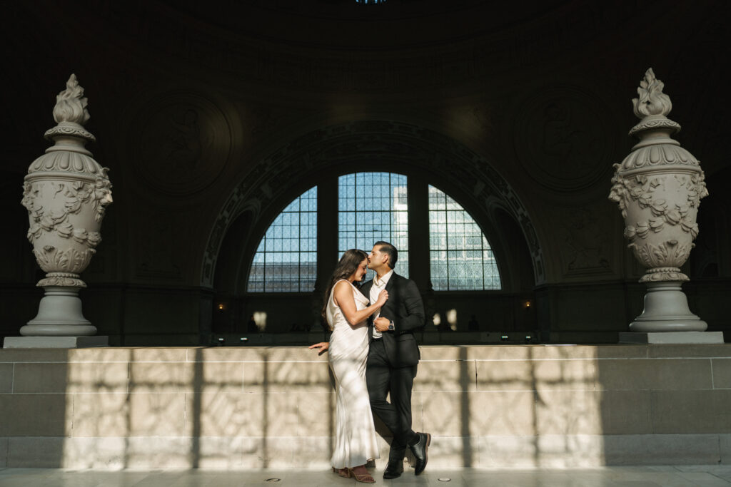 Symmetrical wide shot of newlyweds standing between historic stone urns inside the sunlit San Francisco City Hall.
