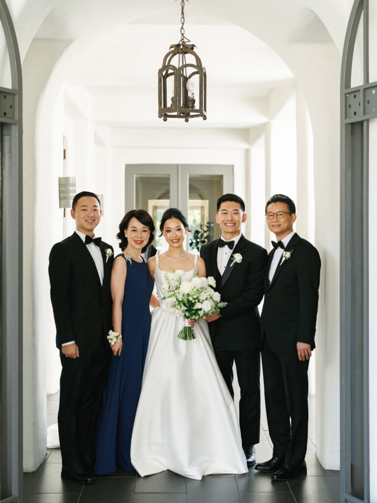 Sophisticated family wedding portrait in a bright arched hallway, showcasing the modern bride with her family dressed in formal black-tie and a navy gown.