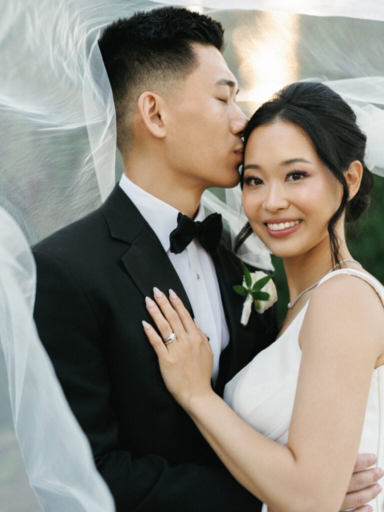 Close-up editorial wedding photography of an elegant Asian bride in a couture gown and groom in a classic tuxedo wrapped in a blowing veil at San Ysidro Ranch.