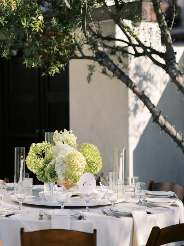 A sophisticated sun-drenched wedding table setting beneath olive trees at a private Southern California estate reception.