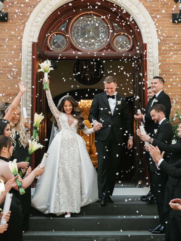 Smiling bride raising a long-stemmed white calla lily bouquet while stepping out of the dark wood arched doors of St. John Cantius Church with her groom under a shower of white paper confetti thrown by wedding guests.