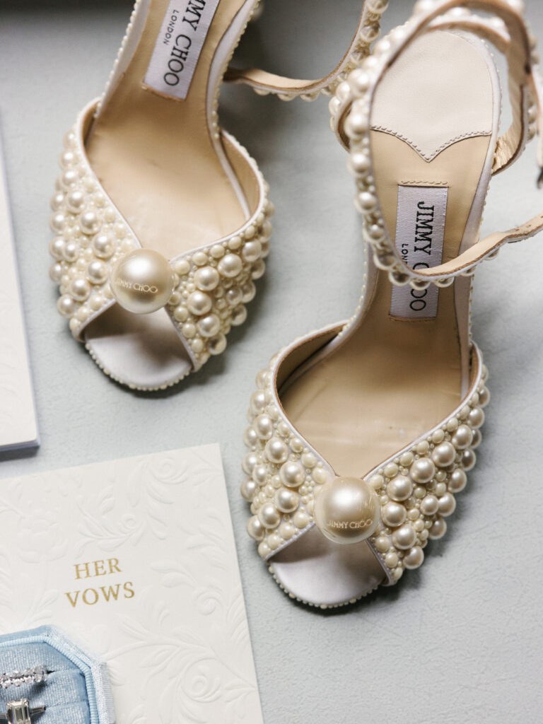 Pearl-embellished Jimmy Choo Sacaria bridal sandals styled on a grey textured surface next to a white Her vows card and light blue ring box for high-end wedding detail photos.