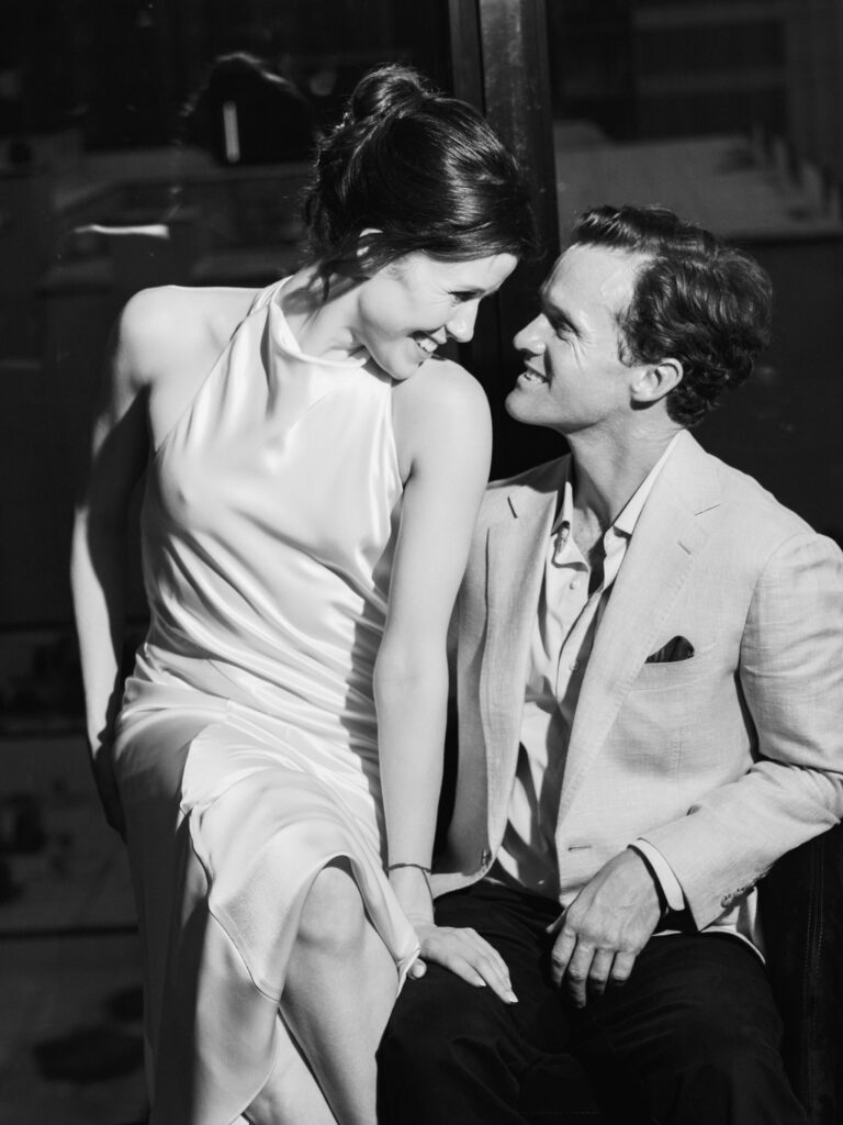 Bride in a sleek satin halter-neck reception dress smiling at her groom in a relaxed tailored summer blazer.