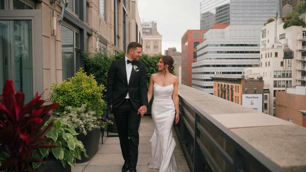A bride and groom walk hand-in-hand on a luxury rooftop terrace with the New York City skyline in the background.