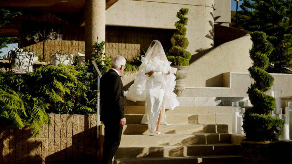 Bride covered by a sheer wedding veil walking down the stone steps of the Pool Terrace at Greystone Mansion toward her father waiting in a tuxedo.