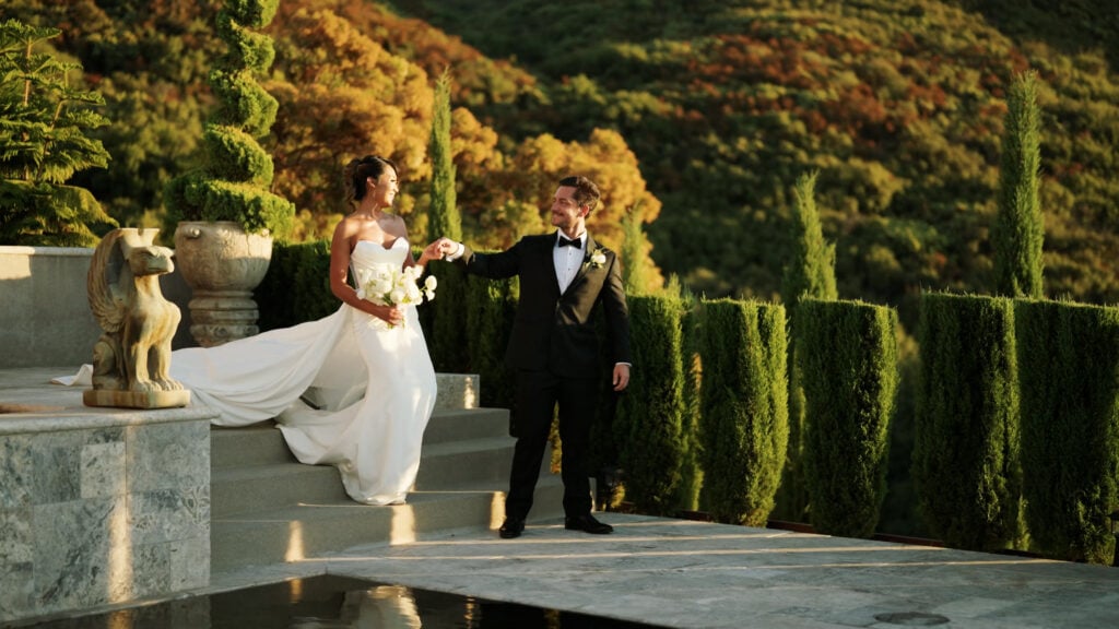 Groom in a black tuxedo holding hands and welcoming his bride down the stone steps at the Pool Terrace of Greystone Mansion, surrounded by cypress trees and a scenic hill background.