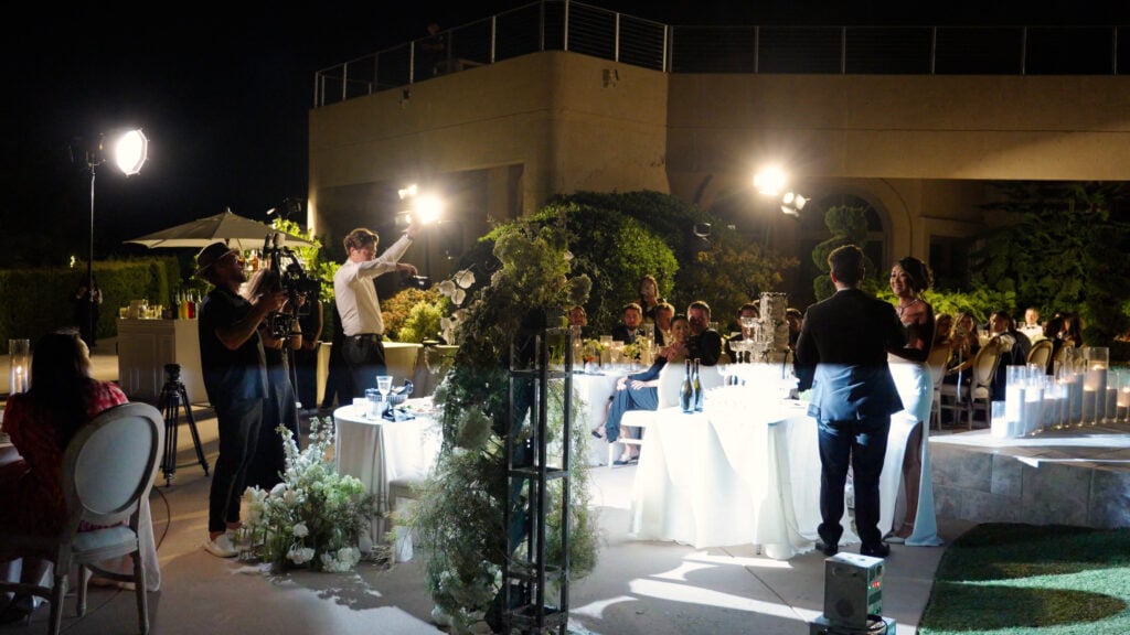 Behind-the-scenes shot of a media crew with camera gimbals and artificial lighting equipment filming a bride and groom facing each other by a candlelit reception table at night at Cielo Farms.