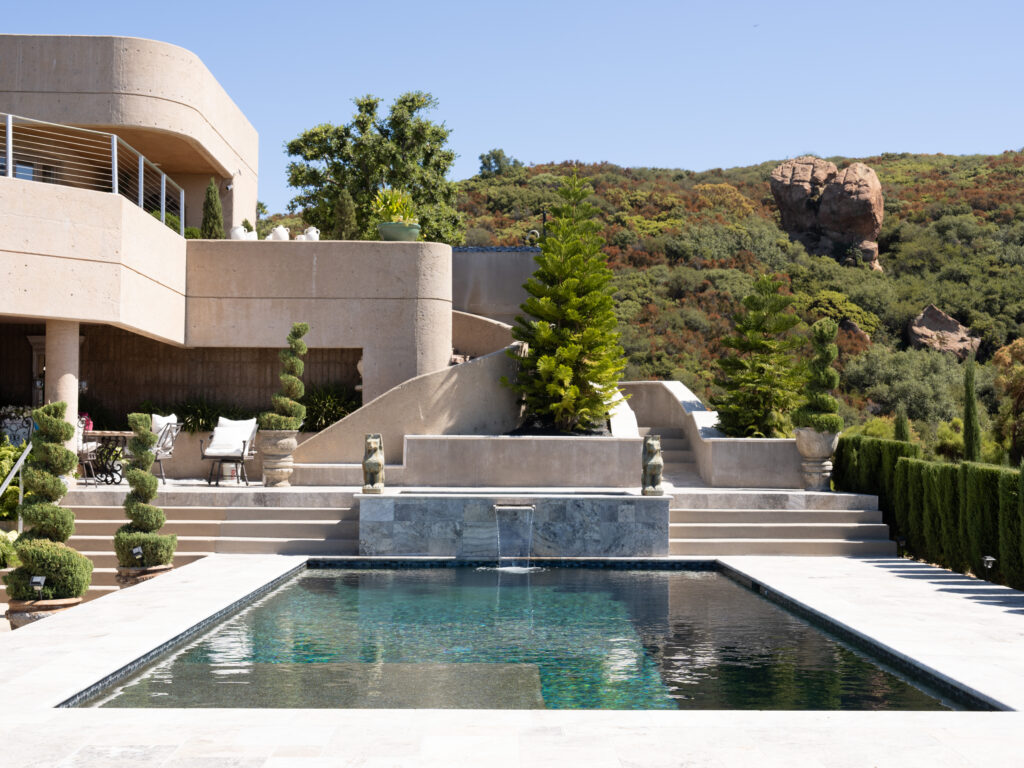 Wide view of a modern estate swimming pool surrounded by stone tiling, spiral topiary trees in pots, green hedges, and patio furniture under a clear blue sky at Greystone Mansion.
