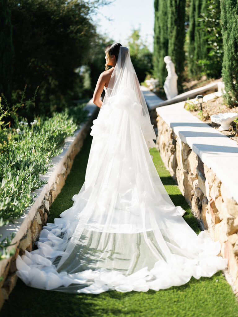 Rear view of a bride showcasing a long cascading tulle dress train and a sheer cathedral veil while standing on a grass path along the Cypress Walk at Greystone Mansion.
