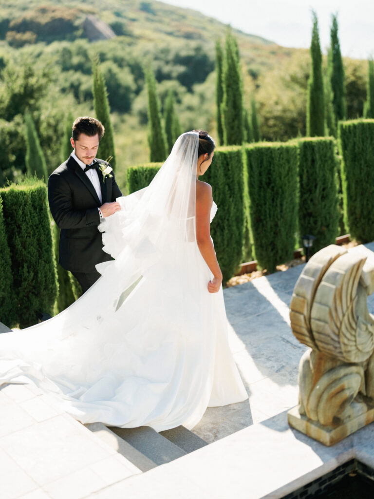Groom in a black tuxedo holding his bride's long dress train as she steps down the stone stairs at the Pool Terrace of Greystone Mansion, next to a historic griffin statue.