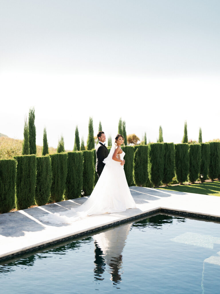 Vertical outdoor portrait of a wedding couple by a pool during a photo and film session at Greystone Mansion estate, highlighting tall cypress trees and a bright sunny sky.