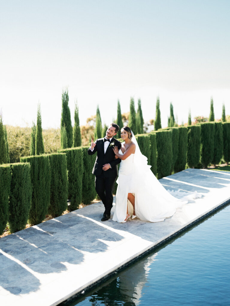 Bride and groom holding arms and walking along the edge of the swimming pool at the Pool Terrace of Greystone Mansion, with the groom gesturing and looking sideways.