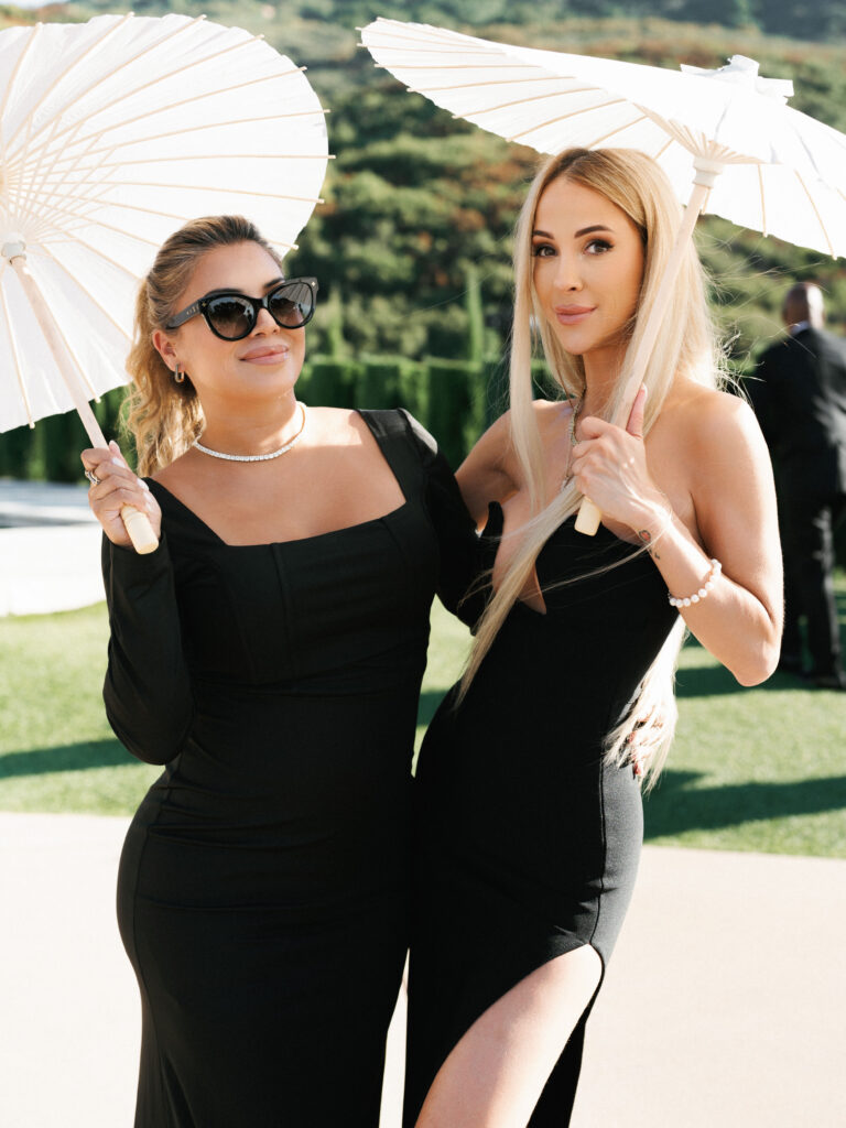 Two women in elegant black evening gowns holding white paper parasols while posing on a sunny lawn during a luxury wedding reception at Greystone Mansion.