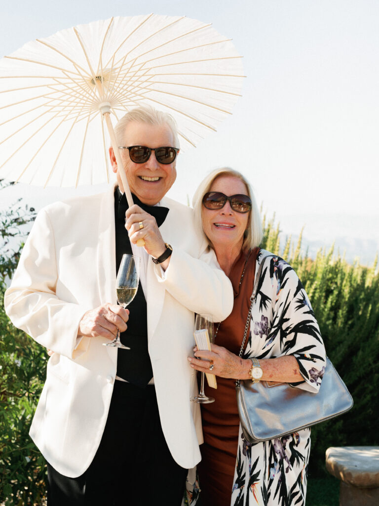 Smiling senior wedding guests in formal attire and sunglasses holding a white paper parasol and a glass of champagne at the Pool Terrace of Greystone Mansion, with green shrubs in the sun.