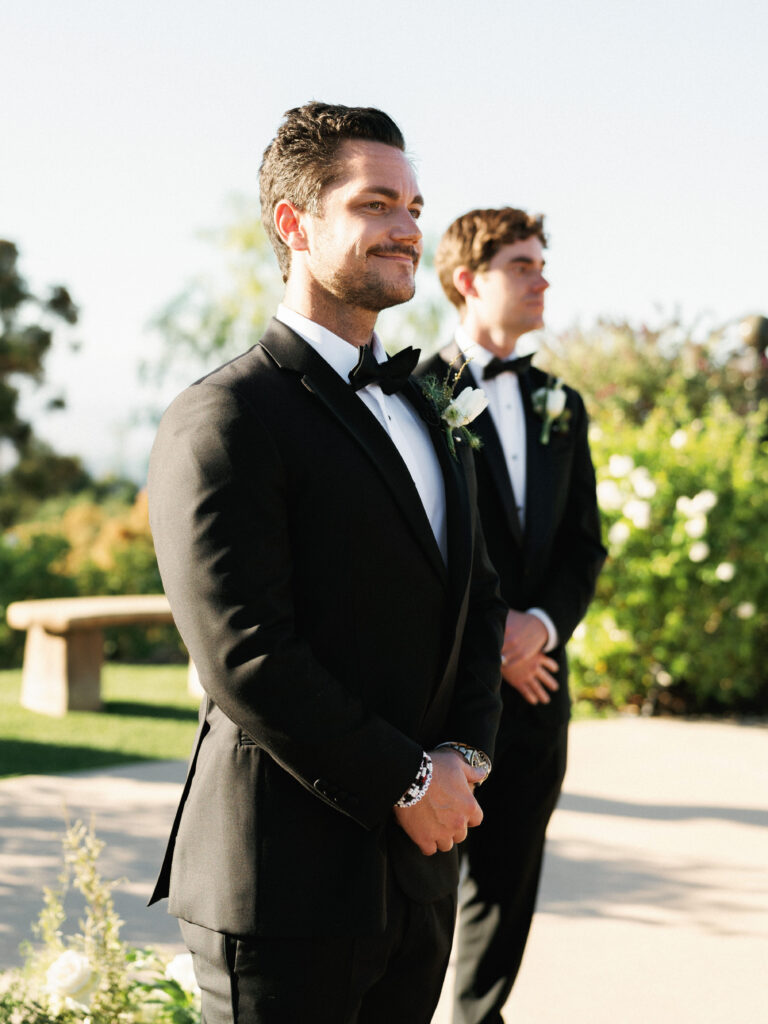 A smiling groom in a black tuxedo with clasped hands standing at the altar during a wedding ceremony at the Formal Gardens of Greystone Mansion, waiting for his bride.