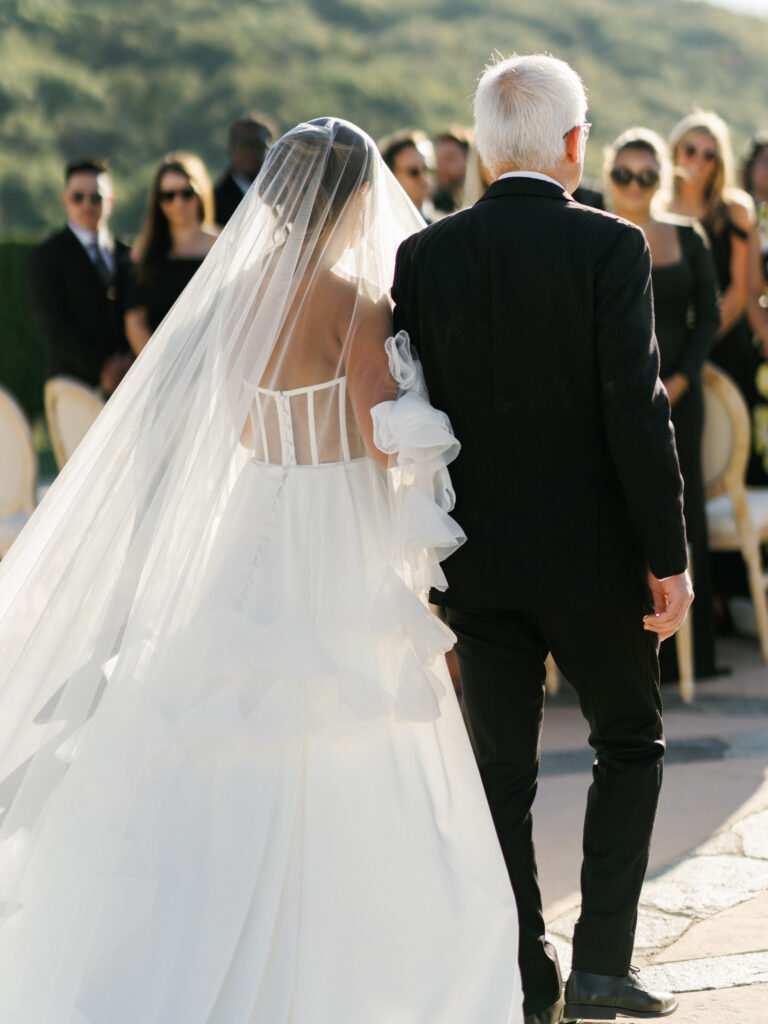 Back view of a bride in a white corset backless wedding dress with ruffle sleeves walking with a man in a black suit past seated guests during an outdoor ceremony.