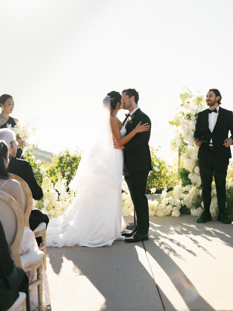 Bride and groom holding each other and kissing at the altar during their wedding ceremony at the Formal Gardens of Greystone Mansion, with an officiant standing nearby.