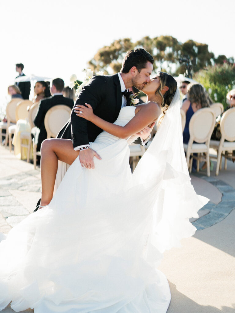 Full-length shot of a groom in a black tuxedo dipping his bride for a kiss on a stone patio, highlighting her white gown with a high leg slit and a flowing veil past applauding guests.